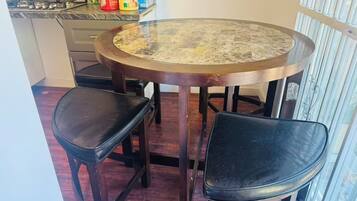 Una mesa de comedor redonda con dos sillas negras, una cocina con estufa a gas y una ventana con persianas verticales.