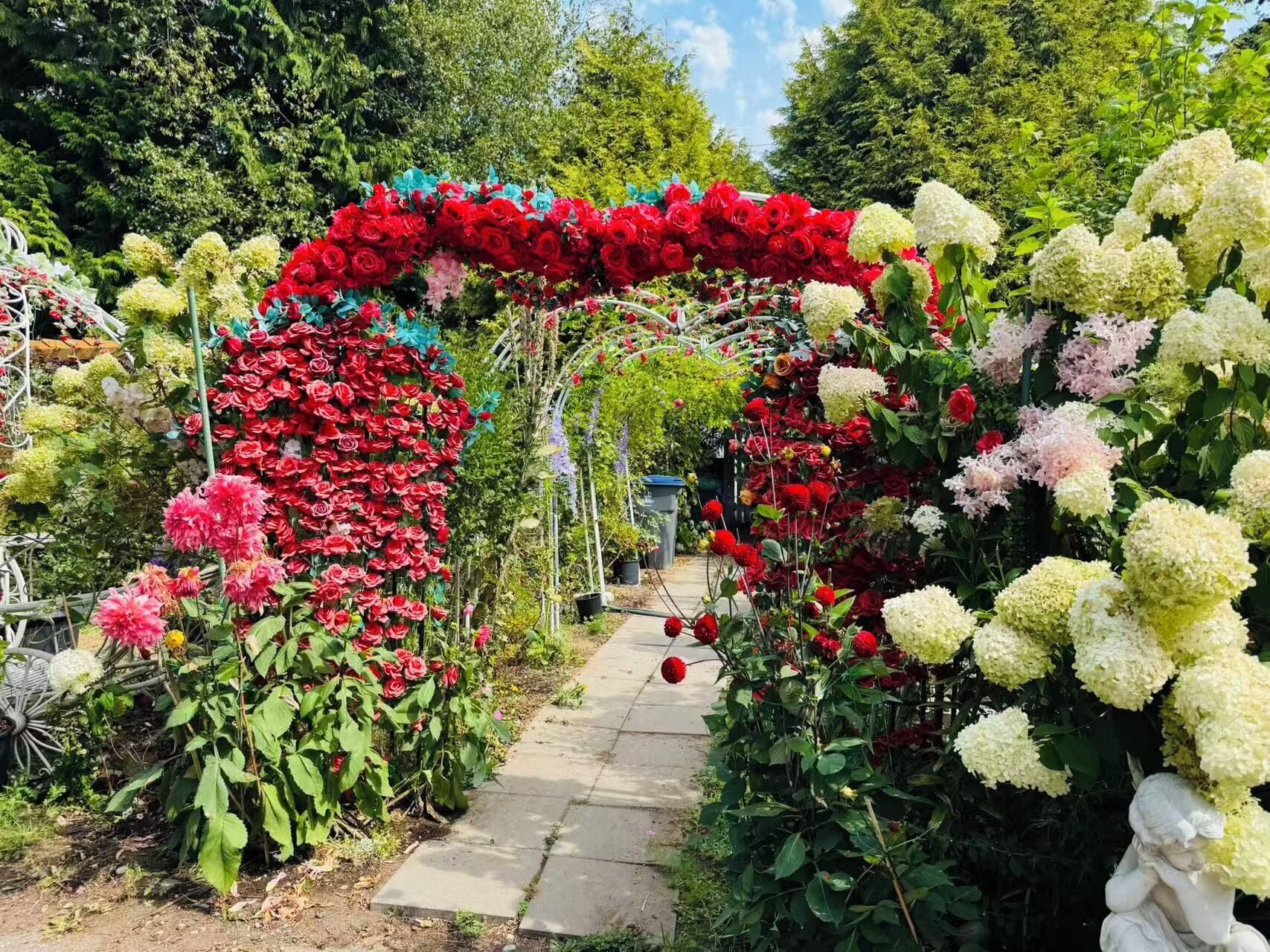 Ein Gartenpfad mit einem blumengeschmückten Bogen, umgeben von verschiedenen Pflanzen und Bäumen.