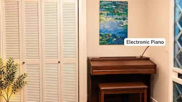 Una habitación con piso de madera, una puerta blanca con paneles verticales, un piano de madera, una araña colgante y un cuadro en la pared.