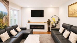 A modern living room with a fireplace, two black leather sofas, a white coffee table, and a flat-screen TV mounted on the wall. - Cozy 3BR Villa Garden View Beach (Surrey)