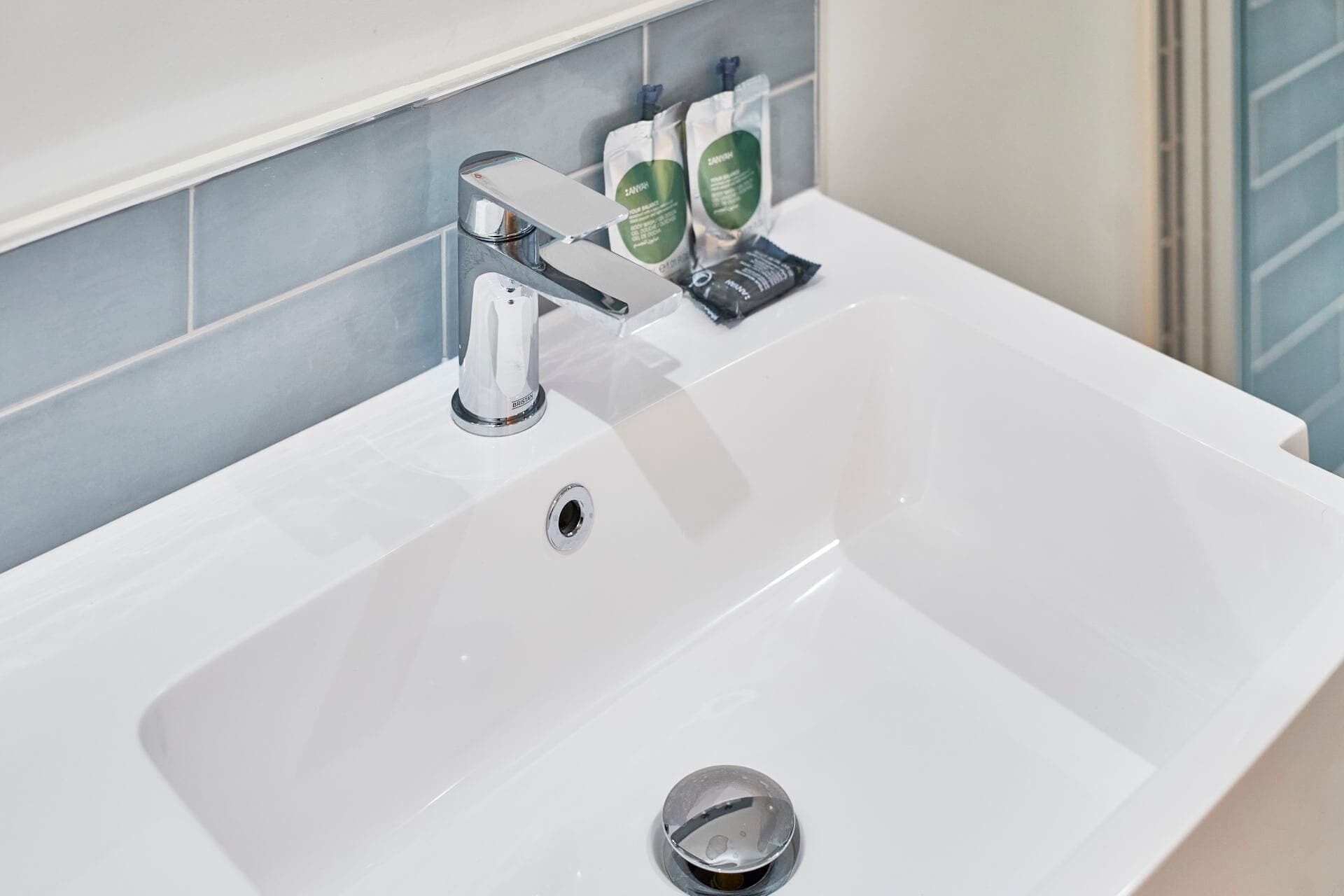 Ein weißes Waschbecken mit Chromarmatur, Ablauf und zwei grünen Flaschen an der Wand.