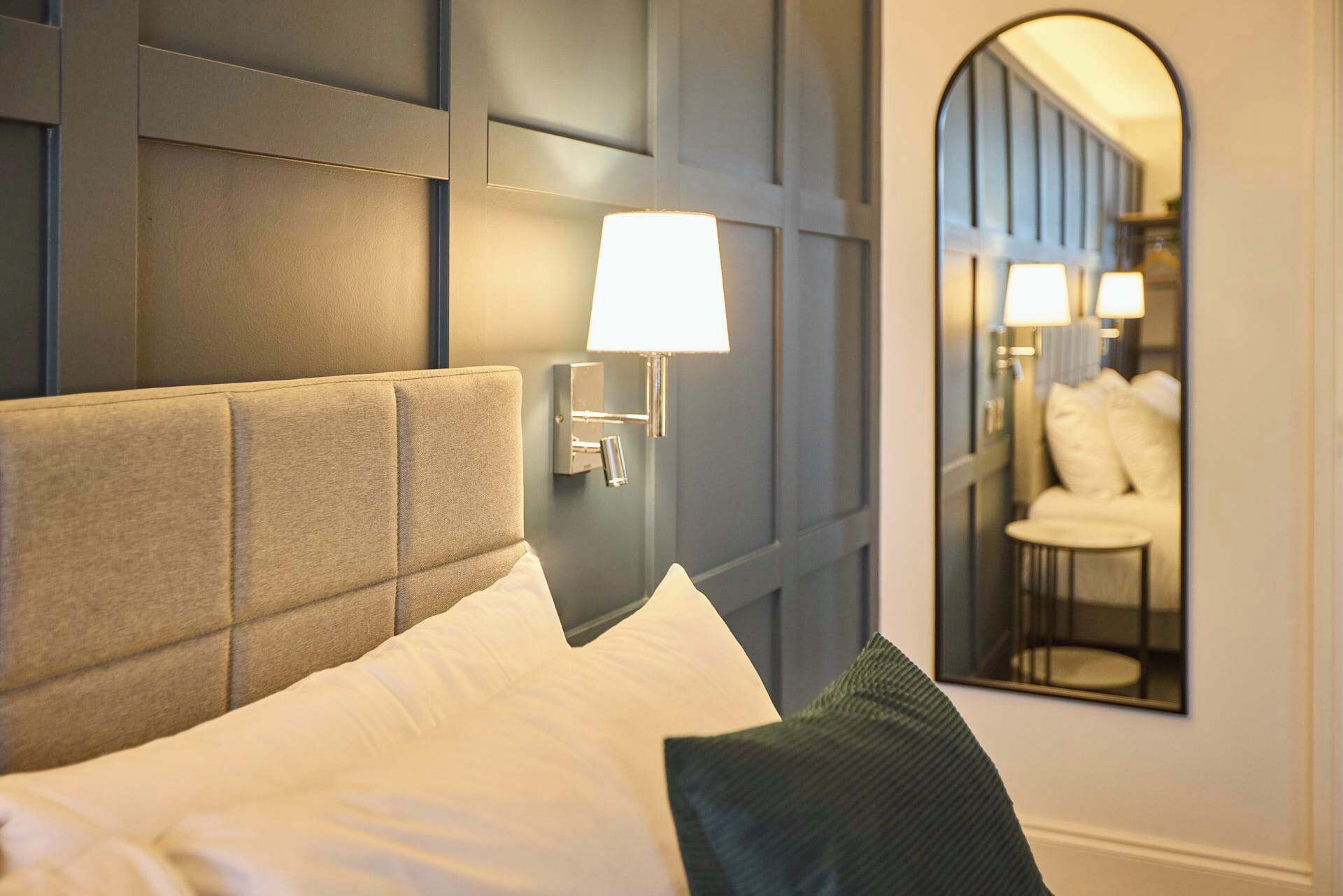 Une chambre d’hôtel avec un grand lit, une tête de lit, une table de chevet, une lampe et un miroir qui reflète un coin salon.