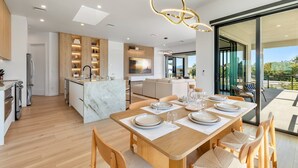 A modern kitchen with wooden cabinets, a marble island, and a dining area with a wooden table set for six. - Upscale Canal-Front Escape w/ Private Pool, Dock & Water Views (Cudjoe Key)