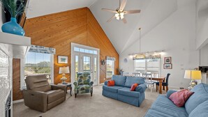 Un salon spacieux avec un haut plafond, des murs en bois et de grandes fenêtres. On y trouve un canapé bleu, un fauteuil en cuir marron et un fauteuil coloré. On aperçoit à l’arrière-plan un coin repas avec une table et des chaises.