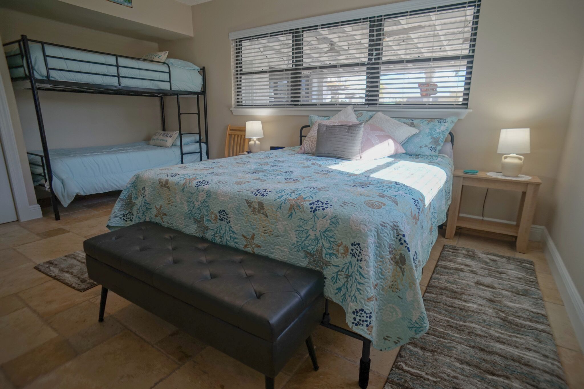 A bedroom with a bunk bed, a large bed with floral bedding, two bedside lamps, a bench, and a window with blinds.