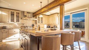 A spacious kitchen with a large island, wooden bar stools, and a view of the outdoors through large windows. - 4BD Fairway Twelve Retreat + Resort Amenities (Sun Valley)