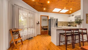 A modern kitchen with wooden flooring, a central island with bar stools, and a view to a living area through a doorway. - The Lilli Pad- A Tranquil Garden Stay (Lilli Pilli)