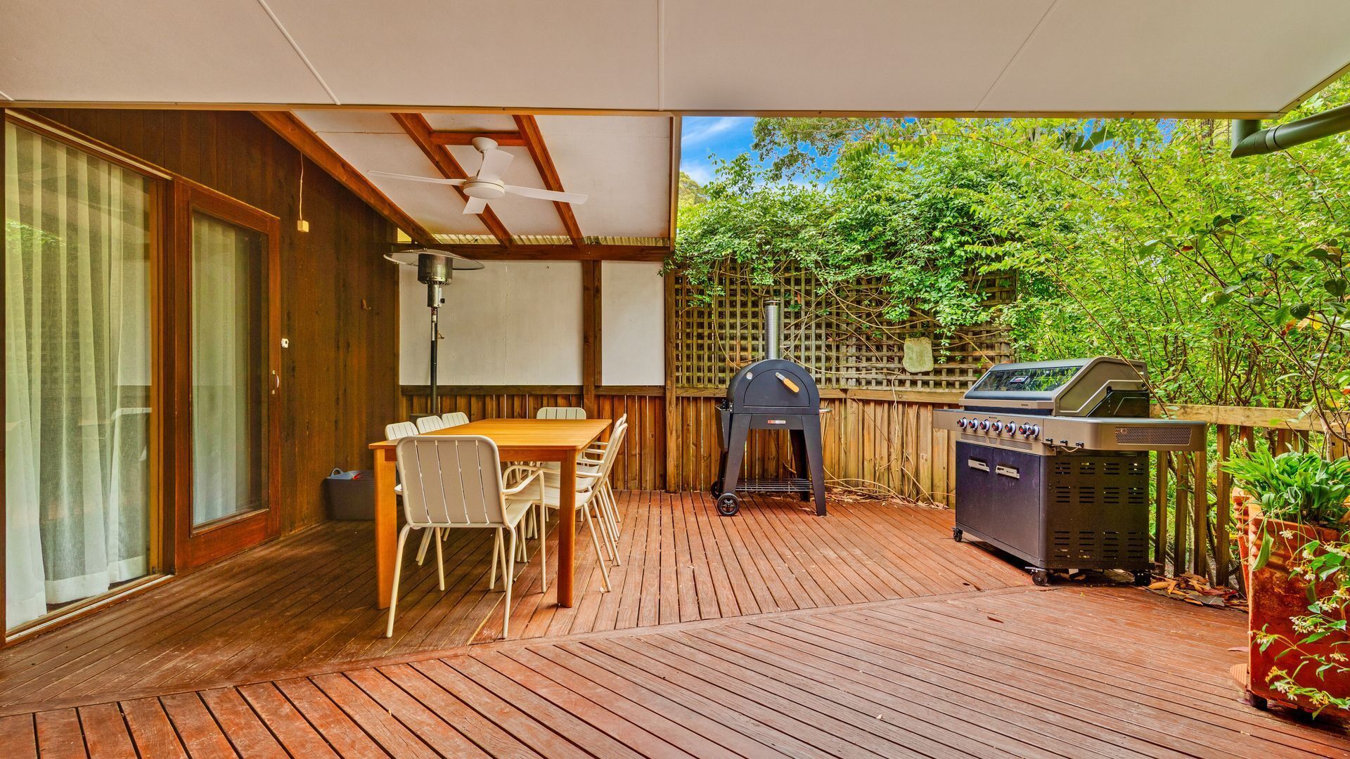 A covered patio with a wooden deck, a table with chairs, a grill, and a ceiling fan.