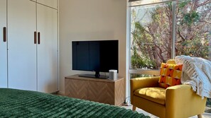 Een slaapkamer met een bed, een televisie op een houten kast, een gele fauteuil en een groot raam met uitzicht op bomen.