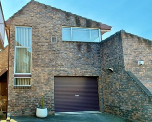Een tweelaags bakstenen huis met een garage deur, een raam met zonwering en een kleine plant in een pot.