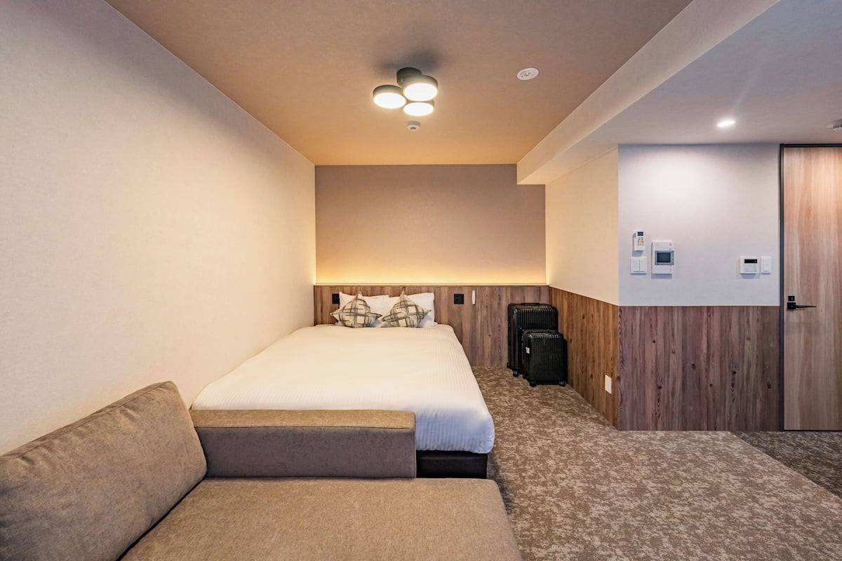 A hotel room with a bed, a sofa, a wooden accent wall, a ceiling light, and a door.