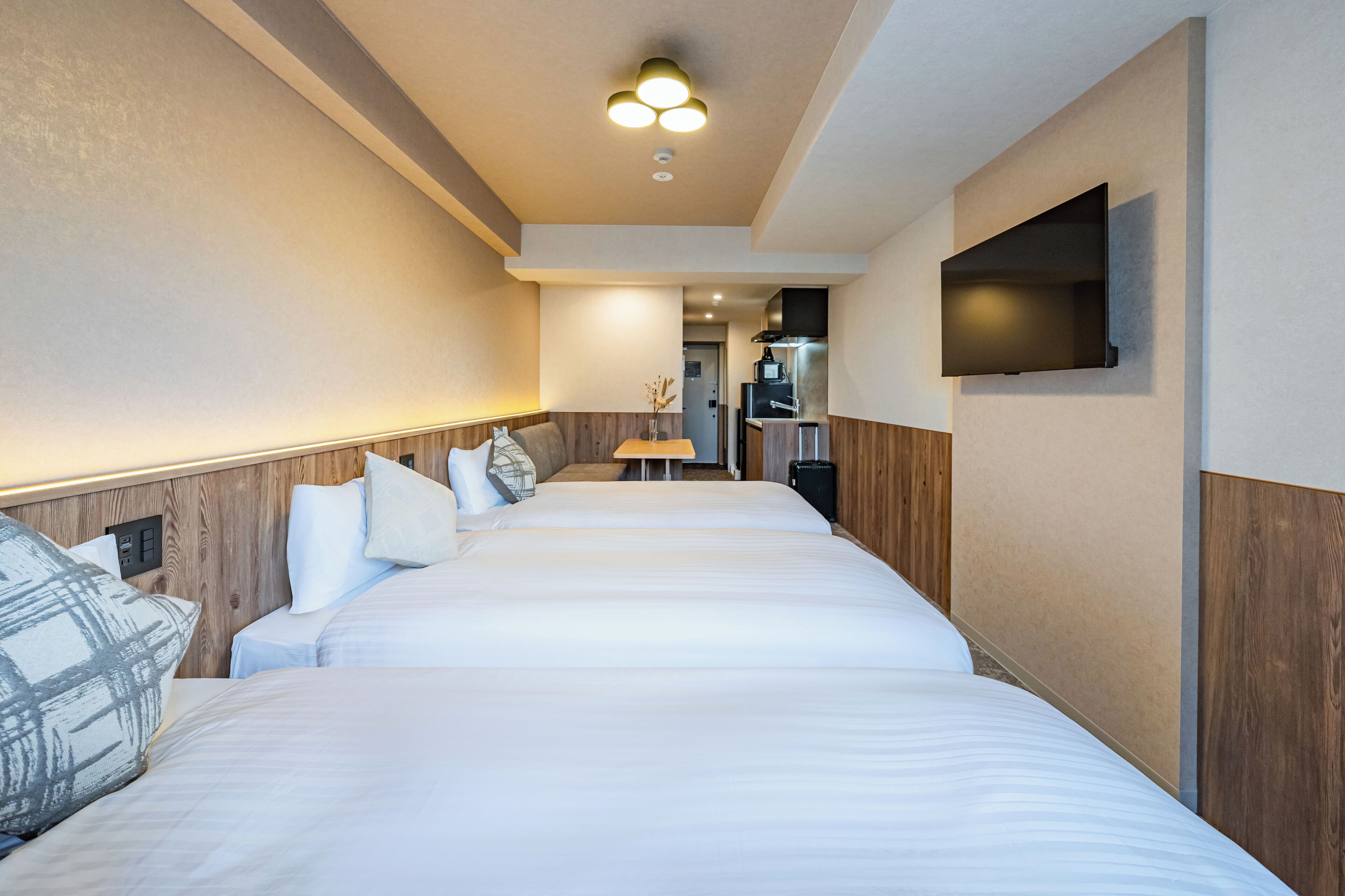 Ein Hotelzimmer mit drei Betten an einer Wand, einem an der Wand befestigten Flachbildfernseher und einem kleinen Tisch mit einer Vase getrockneter Blumen in der Mitte des Raumes.