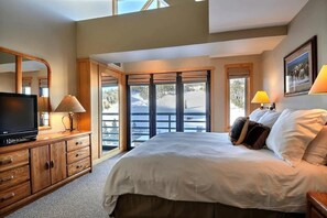 A bedroom with a large bed, wooden dresser, TV, and a view of a snowy landscape through the windows. - Shoshone Condominiums (Gallatin Gateway)