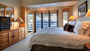 Een slaapkamer met een groot bed, een houten dressoir, een televisie en uitzicht op een besneeuwd landschap door de ramen.