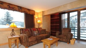 Un salon chaleureux avec un canapé en cuir marron, une table basse en bois et un fauteuil en cuir. Une fenêtre offre une vue sur des montagnes enneigées.