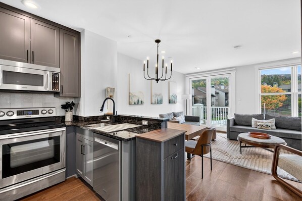 A modern kitchen with stainless steel appliances, a dining area with a wooden table and chairs, and a view of the outdoors through large windows. - Long Trail 2 Bedroom with Stratton Mountain Views (Stratton)