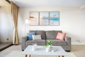 Uma sala de estar moderna com um sofá cinzento, uma mesa de centro branca e três quadros emoldurados com paisagens de praia na parede.