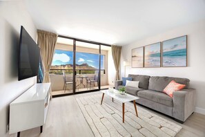 A modern living room with a grey sofa, a flat-screen TV, a coffee table, and a large window with a view of mountains and the sky. - Waikiki Sunset 31st-fl with views, pool & parking (Honolulu)