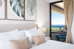 A hotel room with a bed, pillows, a nightstand, a lamp, and a view of the ocean through a window. - Waikiki Sunset 31st-fl with views, pool & parking (Honolulu)