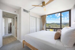 Um quarto com cama, ventoinha no teto, janela com vista para a cidade e porta de acesso a um banheiro.