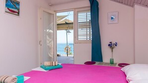 Ein Schlafzimmer mit einem großen Bett, Meerblick durch ein Fenster und einem kleinen Tisch mit Blumen in einer Vase.