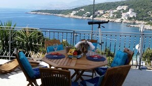 Ein Holztisch mit blauen Kissen, einem Teleskop und einer Obstschale auf einem Balkon mit Blick auf eine Küstenstadt und das Meer.