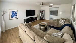 Una sala de estar con un amplio sofá beige, una mesa de centro, un televisor de pantalla plana, una estantería y un ventilador de techo.