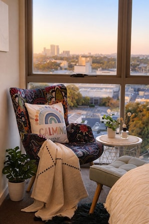 A cozy room with a large window, a colorful chair, a small round table, and a potted plant.