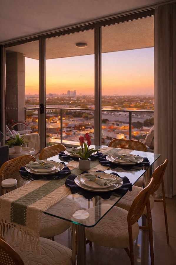 A dining area with a glass table, wicker chairs, and a view of a cityscape at sunset.