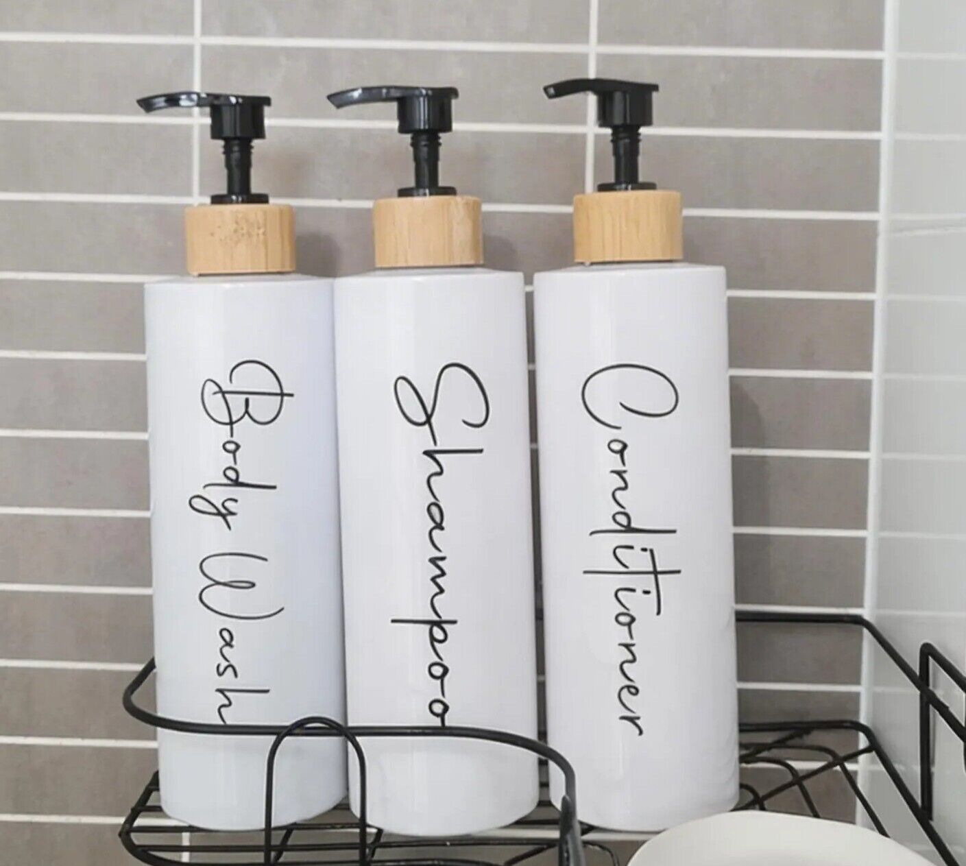 Three white bottles with black pumps and wooden caps labeled "Body Wash," "Shampoo," and "Conditioner" on a metal rack.