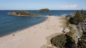 A beach with a volleyball court, a parking area, and a small island in the background. - Sunrise & Sea views - Luxe downtown with 2 carpark (Mount Maunganui)