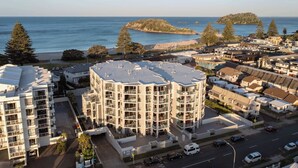 A coastal residential area with modern apartment buildings, a beach, and a small island in the background. - Sunrise & Sea views - Luxe downtown with 2 carpark (Mount Maunganui)