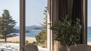 A room with a large window overlooking a beach, a potted plant, and a dining area with chairs and a table. - Sunrise & Sea views - Luxe downtown with 2 carpark (Mount Maunganui)