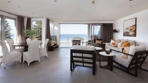 A spacious living room with a large window offering a view of the sea, a sofa set, a dining table with chairs, and a small side table. - Sunrise & Sea views - Luxe downtown with 2 carpark (Mount Maunganui)