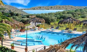A pool area with lounge chairs and umbrellas, surrounded by greenery and hills. - Villa Galini Sifis (Apokoronas)