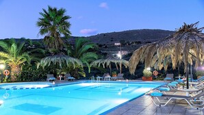 Μια πισίνα με ξαπλώστρες και ομπρέλες, περιτριγυρισμένη από φοίνικες και με ένα επιβλητικό ορεινό τοπίο στο βάθος.