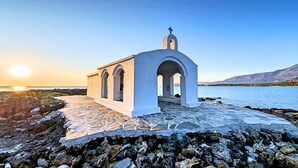 A white chapel with a cross on top, located on a rocky shore with a view of the sea and mountains in the background. - Villa Galini Sifis (Apokoronas)