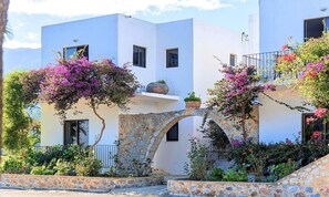 Un edificio bianco con archi in pietra e balconi, immerso nel verde e circondato da piante fiorite.