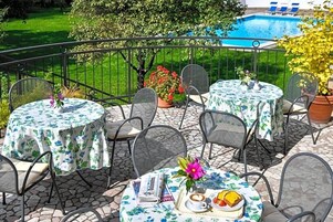 A terrace with round tables and chairs, a pool in the background, and potted plants.