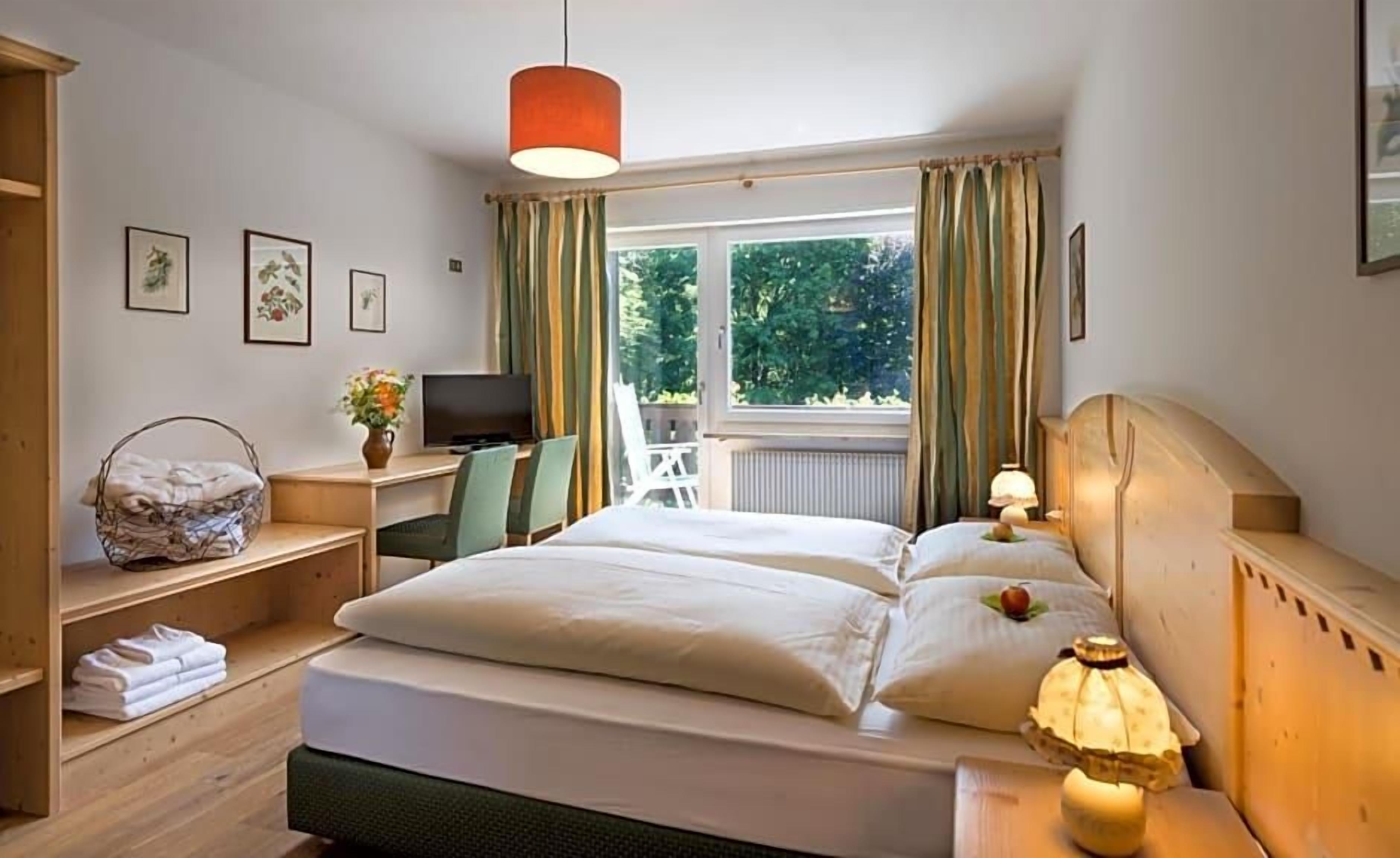 Ein Hotelzimmer mit einem Bett, Nachttischlampen, einem Schreibtisch mit Computer, einem Sessel, einem Fenster mit Vorhängen und Blick ins Grüne.