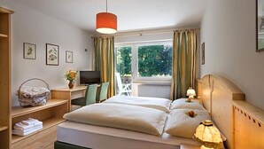 A hotel room with a bed, bedside lamps, a desk with a computer, a chair, a window with curtains, and a view of greenery outside. - Perwanger (Fie allo Sciliar)