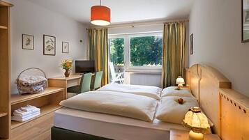 Ein Hotelzimmer mit einem Bett, Nachttischlampen, einem Schreibtisch mit Computer, einem Sessel, einem Fenster mit Vorhängen und Blick ins Grüne.