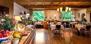 A dining area with wooden tables and chairs, a buffet with fruits and pastries, and a view of greenery through large windows. - Perwanger (Fie allo Sciliar)