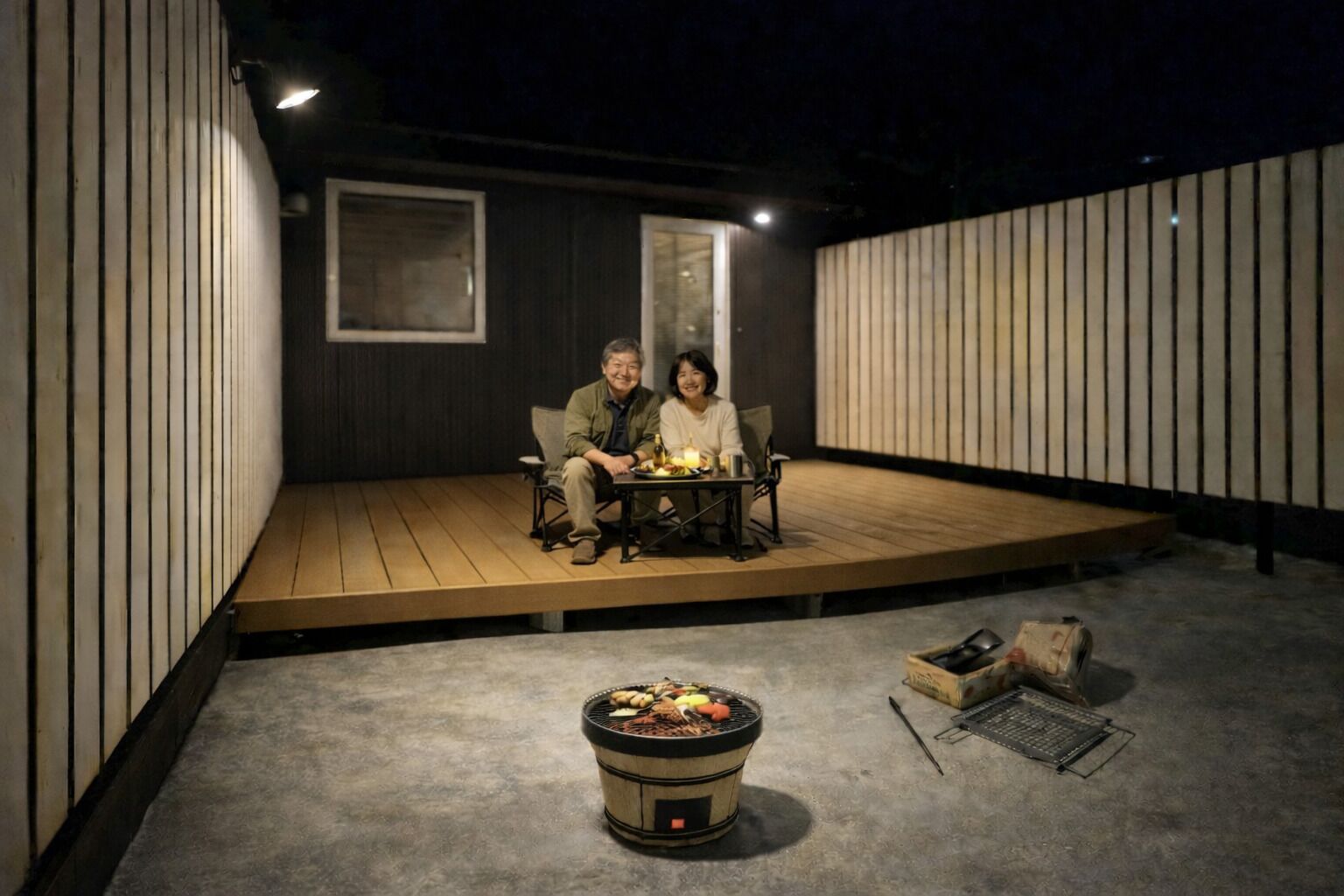 A couple seated at a table on a wooden deck with a grill in the foreground.