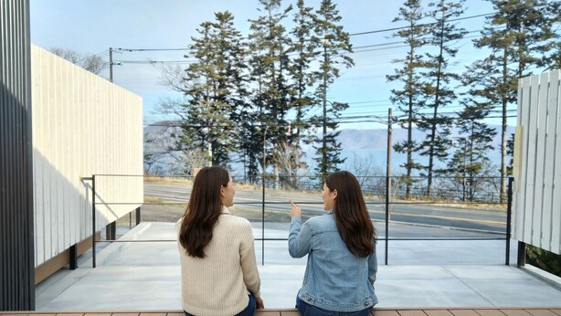 Two people sitting on a wooden deck overlooking a road with trees and a building in the background. - ATTA Toya  - Lake Resort (Sobetsu)