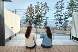 Two people sitting on a wooden deck overlooking a road with trees and a building in the background. - ATTA Toya  - Lake Resort (Sobetsu)