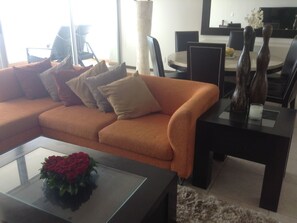 Ein modernes Wohnzimmer mit einer Eckcouch, einem Glastisch mit Blumenarrangement und einem Essbereich mit rundem Tisch und Stühlen.