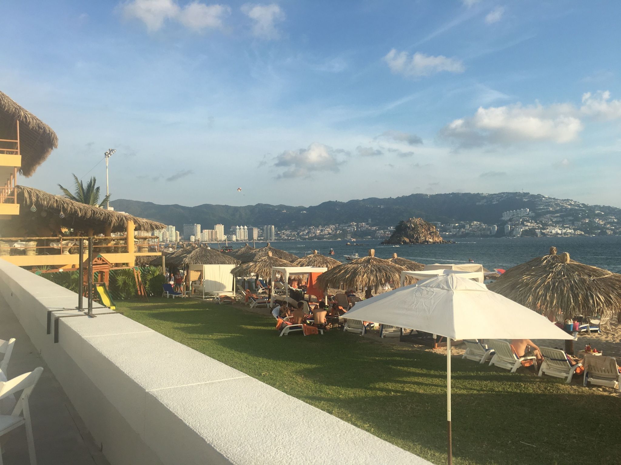 Vista sulla spiaggia con ombrelloni di paglia, sdraio e sullo sfondo mare e edifici distanti.