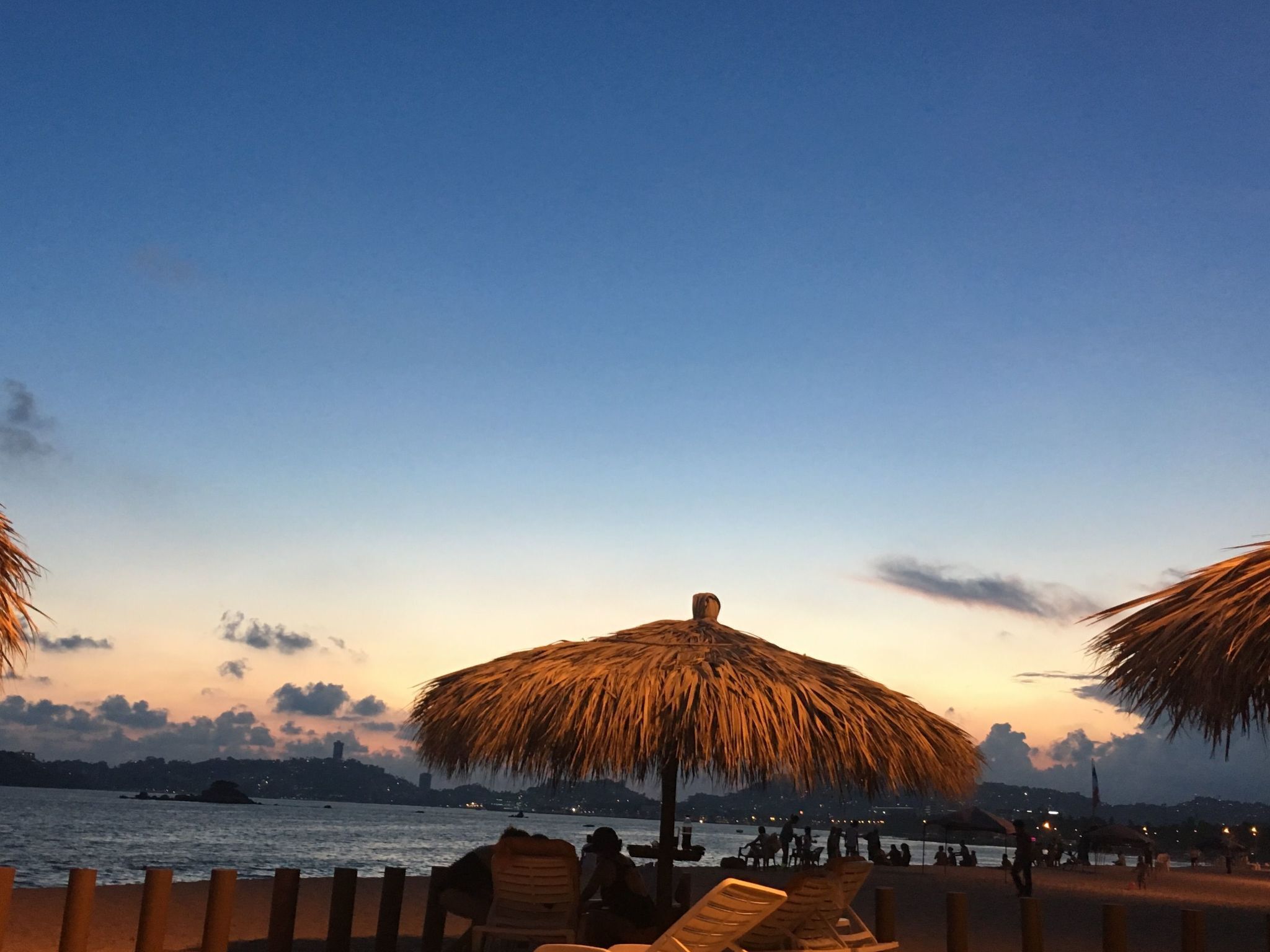 A beach with thatched umbrellas and lounge chairs, a calm sea, and a city skyline in the distance.