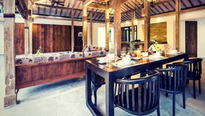 A rustic dining area with a long wooden table set for four, surrounded by dark chairs. A bench with patterned cushions is against the wall.